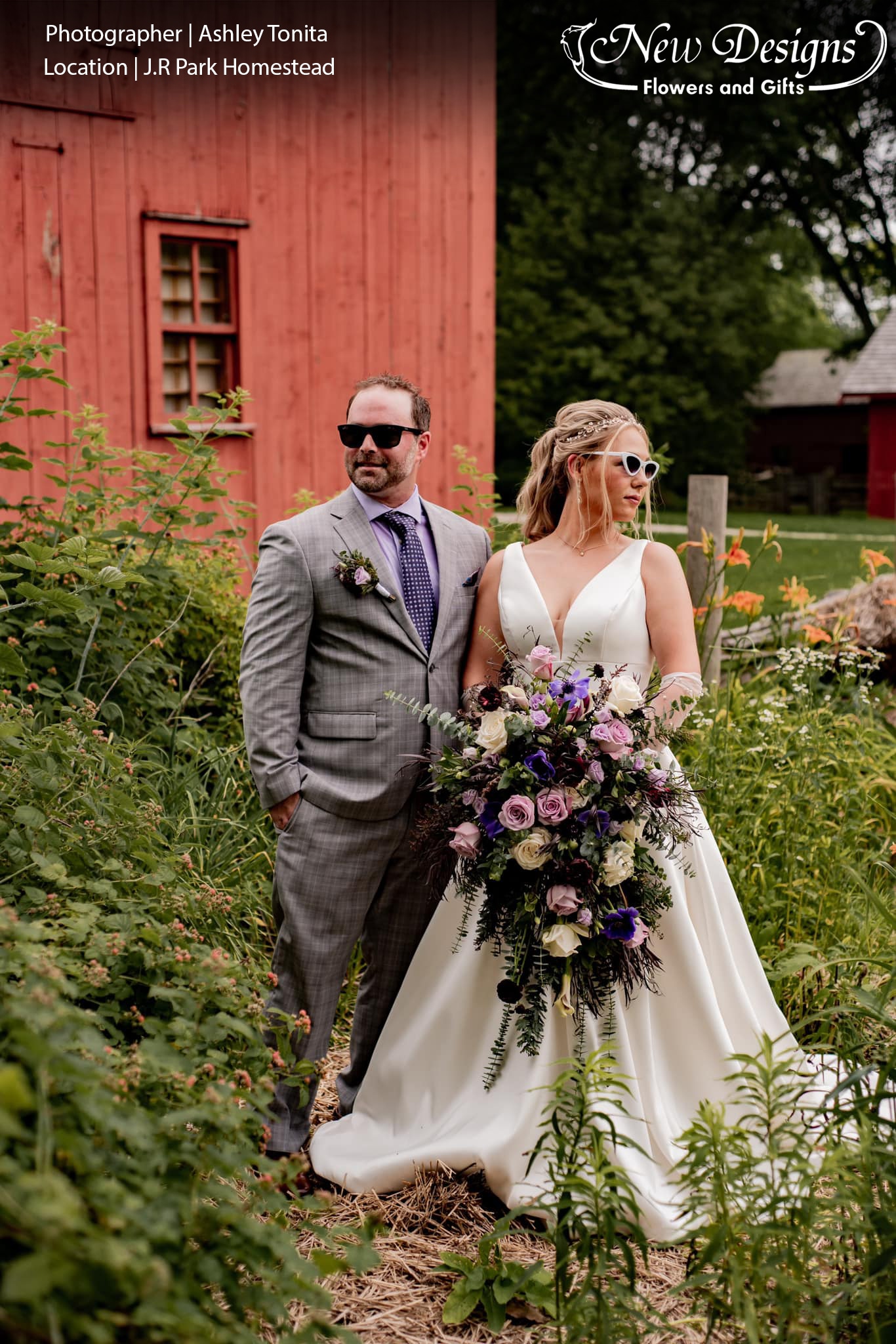 A bride and Groom stand in an old homestead, against a red barn, and Overflowing wildflower garden, The bride holds an overflowing cascading bouquet in lush greens and various shades of bright and deep Purple, Green and White flowers